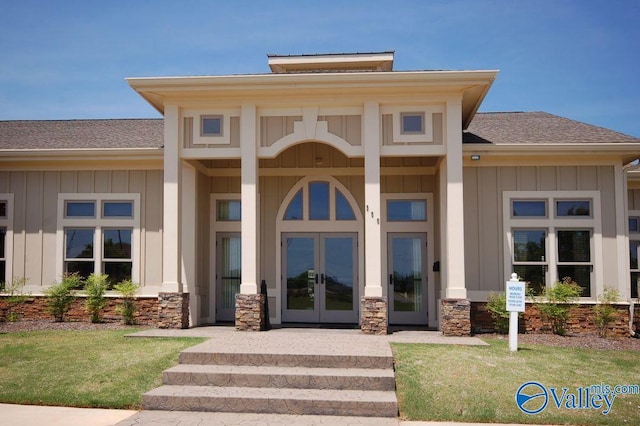 property entrance with board and batten siding, french doors, roof with shingles, a lawn, and stone siding
