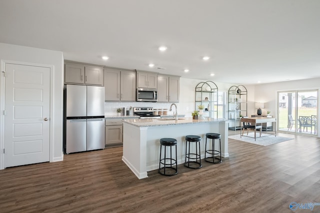 kitchen featuring dark wood-type flooring, gray cabinetry, light stone counters, an island with sink, and stainless steel appliances