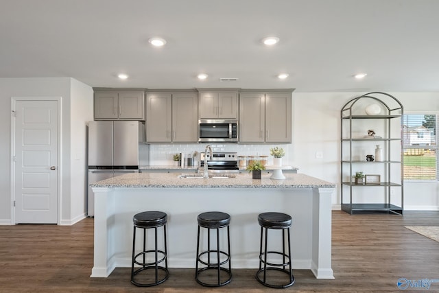 kitchen featuring stainless steel appliances, gray cabinets, an island with sink, and light stone countertops