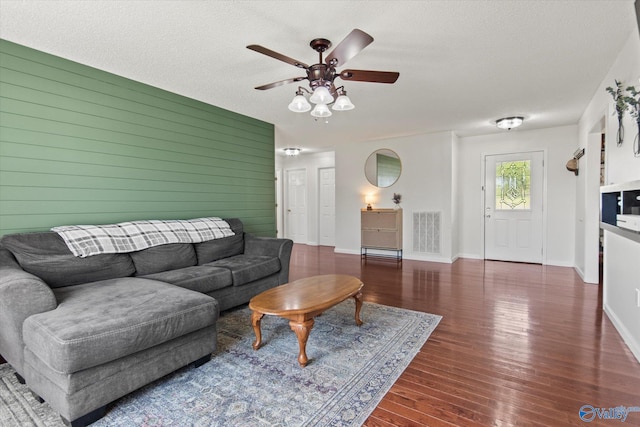 living room with a textured ceiling, visible vents, wooden walls, ceiling fan, and wood finished floors