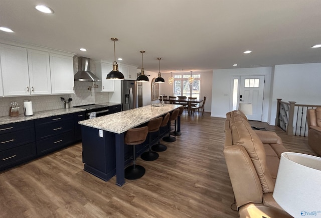 kitchen featuring white cabinets, hanging light fixtures, a center island with sink, stainless steel refrigerator with ice dispenser, and wall chimney range hood