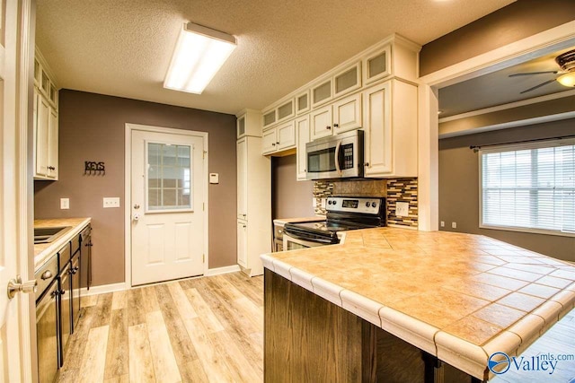 kitchen with decorative backsplash, tile countertops, stainless steel appliances, light wood finished floors, and glass insert cabinets