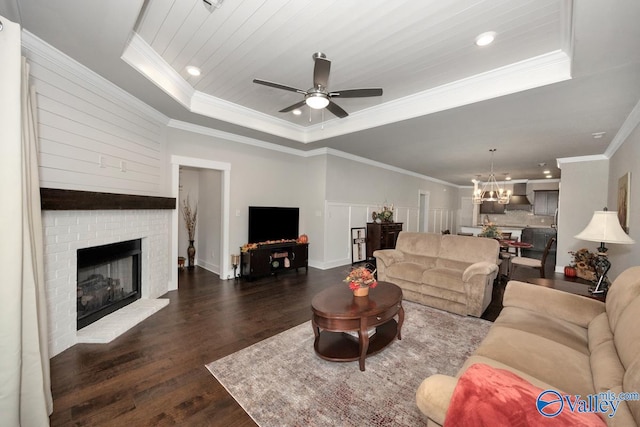 living room with a tray ceiling, ornamental molding, dark wood-type flooring, ceiling fan with notable chandelier, and a fireplace