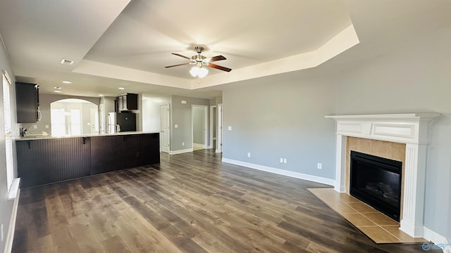 unfurnished living room with a tray ceiling, dark wood-type flooring, a tile fireplace, and ceiling fan