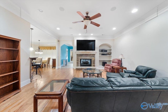 living room featuring built in shelves, a stone fireplace, ceiling fan, light hardwood / wood-style floors, and ornamental molding