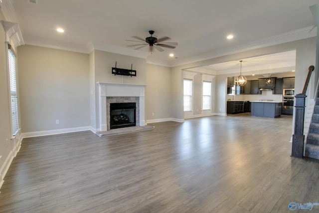 unfurnished living room featuring ceiling fan, a stone fireplace, wood finished floors, stairs, and crown molding