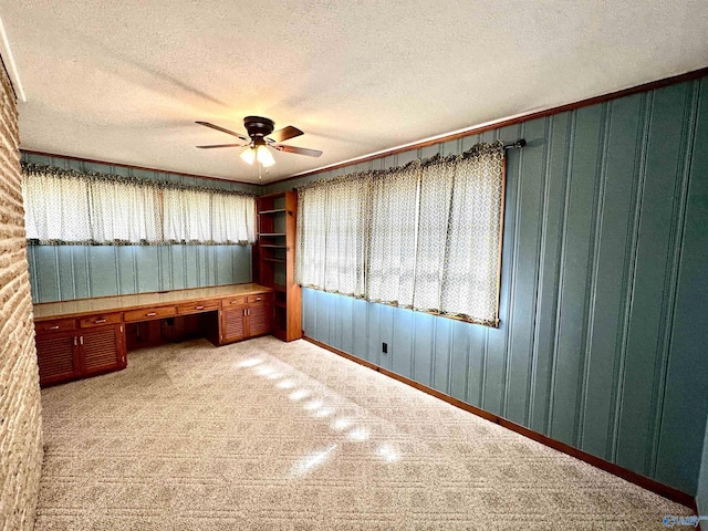 unfurnished office featuring wooden walls, a ceiling fan, light colored carpet, a textured ceiling, and built in desk