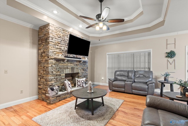 living room with a stone fireplace, wood-type flooring, ornamental molding, ceiling fan, and a tray ceiling