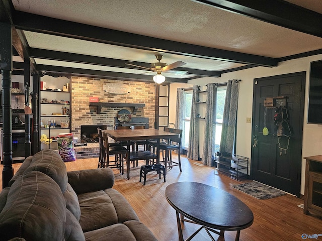 living room featuring ceiling fan, a brick fireplace, a textured ceiling, beam ceiling, and light hardwood / wood-style flooring