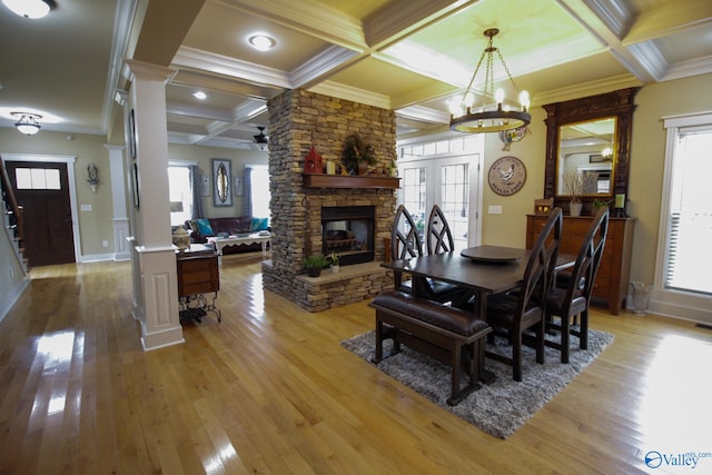 dining room featuring a stone fireplace, light hardwood / wood-style floors, ceiling fan with notable chandelier, ornate columns, and coffered ceiling