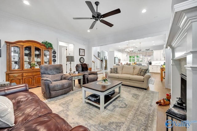living room featuring crown molding, a fireplace, stairway, ceiling fan with notable chandelier, and light wood-style flooring