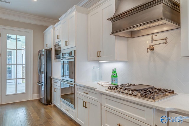 kitchen with ornamental molding, white cabinets, custom exhaust hood, appliances with stainless steel finishes, and light hardwood / wood-style floors