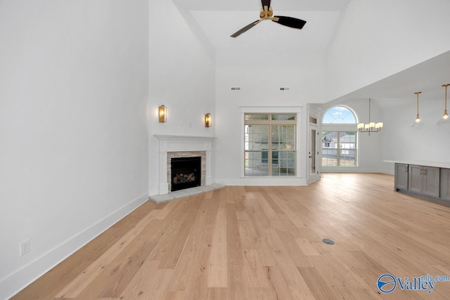 unfurnished living room featuring light wood-type flooring, baseboards, a fireplace with raised hearth, ceiling fan with notable chandelier, and high vaulted ceiling