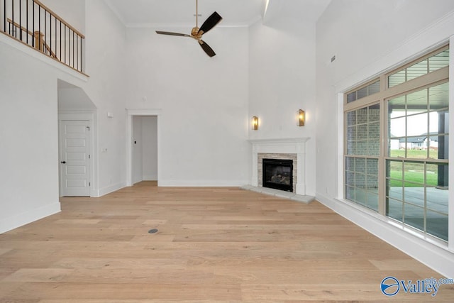 unfurnished living room featuring a stone fireplace, ceiling fan, light wood finished floors, baseboards, and a high ceiling