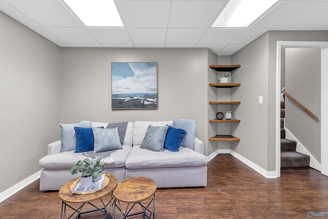 living room featuring built in shelves, wood finished floors, baseboards, a drop ceiling, and stairs