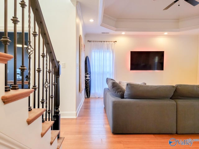 living room with wood finished floors, a ceiling fan, stairs, a raised ceiling, and crown molding