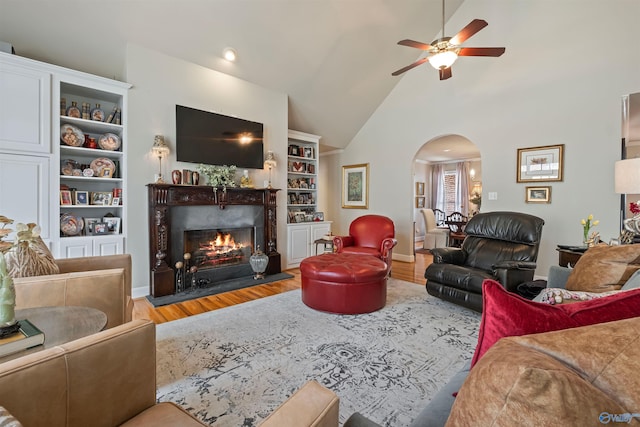 living area featuring high vaulted ceiling, a ceiling fan, wood finished floors, arched walkways, and a lit fireplace