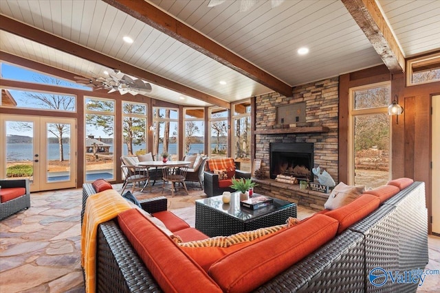 living room featuring vaulted ceiling with beams, a stone fireplace, a water view, a ceiling fan, and stone tile flooring