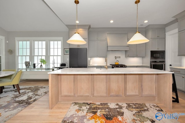 kitchen with an island with sink, premium range hood, black appliances, gray cabinets, and light wood-type flooring