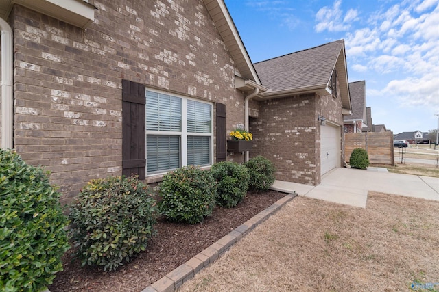 view of side of home with driveway, fence, brick siding, a shingled roof, and a garage