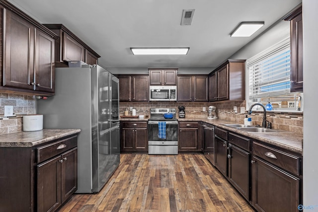 kitchen with a sink, dark wood-type flooring, tasteful backsplash, dark brown cabinetry, and appliances with stainless steel finishes