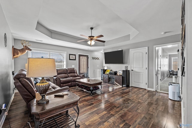 living room with plenty of natural light, a ceiling fan, dark wood-style flooring, and a tray ceiling