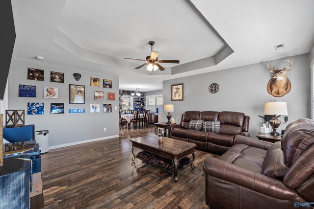 living room with a tray ceiling, visible vents, baseboards, wood finished floors, and ceiling fan with notable chandelier