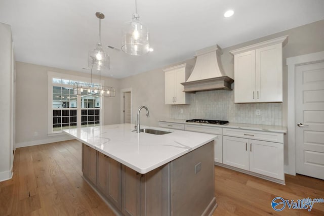 kitchen featuring a center island with sink, a sink, gas cooktop, white cabinets, and custom exhaust hood