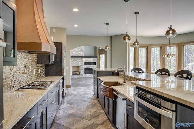 kitchen with tile patterned flooring, custom exhaust hood, appliances with stainless steel finishes, a stone fireplace, and decorative light fixtures