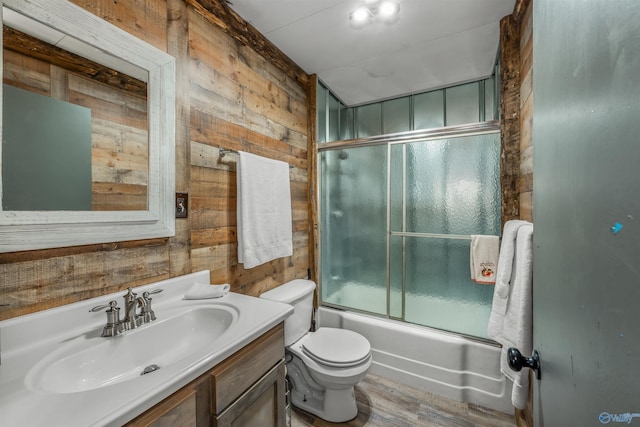 full bathroom with bath / shower combo with glass door, wood walls, wood-type flooring, vanity, and toilet