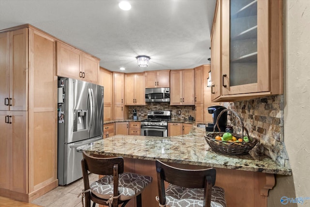 kitchen featuring light stone countertops, light brown cabinets, stainless steel appliances, kitchen peninsula, and decorative backsplash