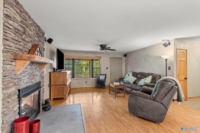 living room with ceiling fan, light hardwood / wood-style floors, a stone fireplace, and a textured ceiling