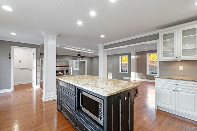 kitchen with white cabinetry, stainless steel microwave, ceiling fan, a stone fireplace, and wood-type flooring