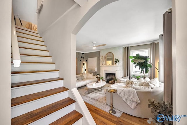 living area with wood finished floors, stairs, arched walkways, a large fireplace, and ceiling fan