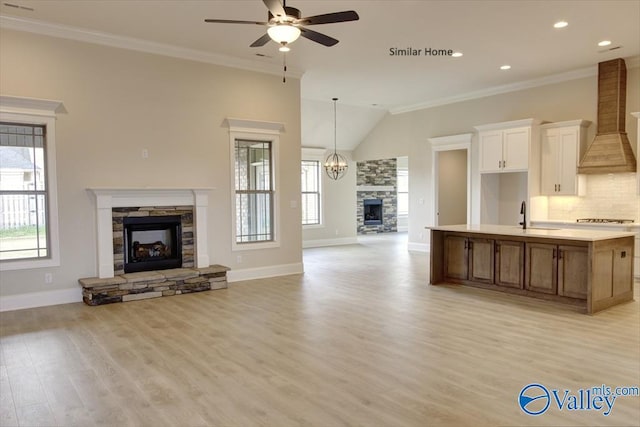 kitchen with ceiling fan with notable chandelier, ornamental molding, custom range hood, an island with sink, and a stone fireplace