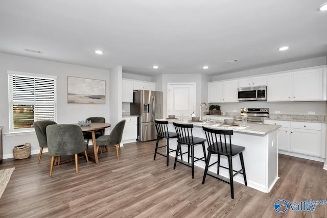 kitchen with white cabinetry, wood-type flooring, stainless steel appliances, light stone countertops, and a center island with sink