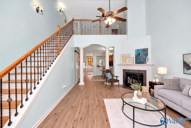 living room with ceiling fan, light hardwood / wood-style floors, ornate columns, a towering ceiling, and a tiled fireplace