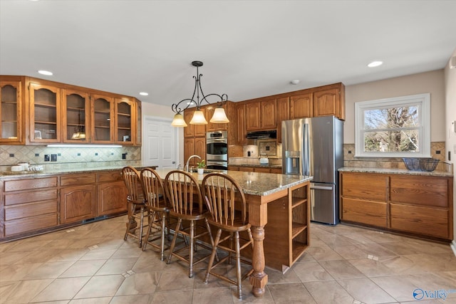 kitchen with light stone countertops, an island with sink, brown cabinetry, and appliances with stainless steel finishes