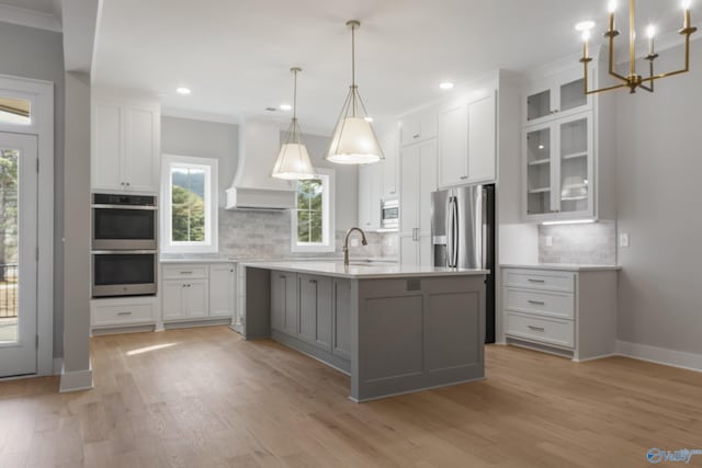 kitchen featuring appliances with stainless steel finishes, white cabinets, custom exhaust hood, hanging light fixtures, and a kitchen island with sink