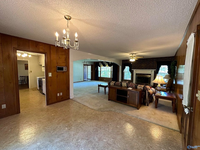 carpeted living room with wooden walls, a textured ceiling, ceiling fan with notable chandelier, a brick fireplace, and brick wall