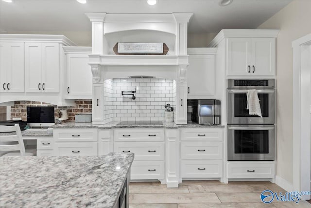 kitchen with double oven, tasteful backsplash, light stone countertops, and white cabinets