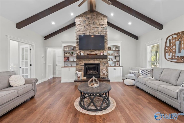 living room featuring ceiling fan, a stone fireplace, wood finished floors, baseboards, and beam ceiling