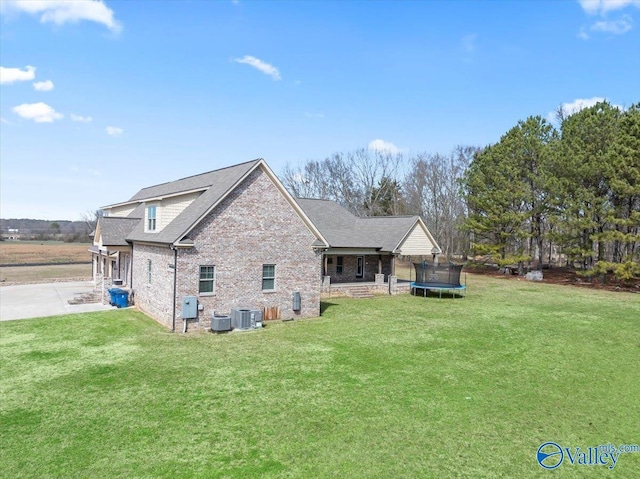 rear view of property featuring cooling unit, brick siding, roof with shingles, a lawn, and a trampoline