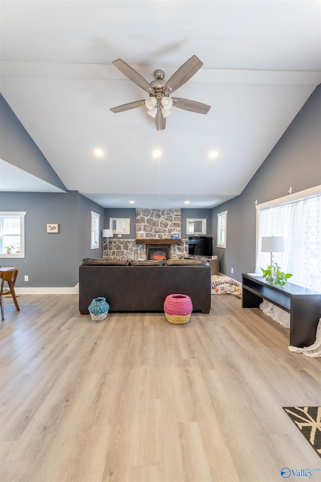 living room with ceiling fan, a stone fireplace, vaulted ceiling, and light wood-type flooring