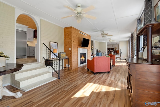 living room with stairway, a ceiling fan, a glass covered fireplace, light wood-style floors, and brick wall