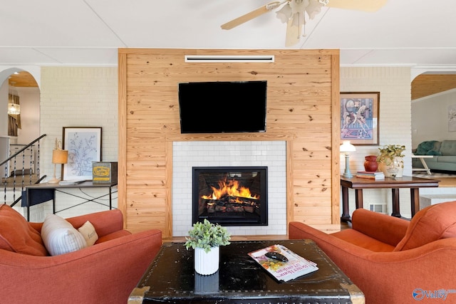 living area with wood walls, arched walkways, crown molding, a glass covered fireplace, and ceiling fan