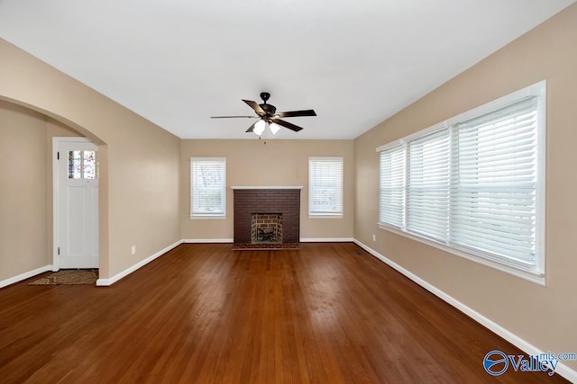 unfurnished living room with arched walkways, a ceiling fan, a brick fireplace, wood finished floors, and baseboards