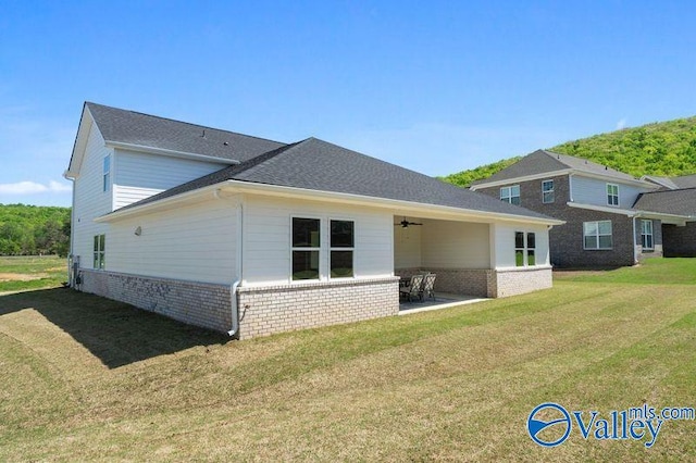 back of property with roof with shingles, a ceiling fan, a patio area, brick siding, and a lawn