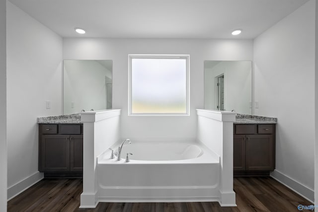 bathroom with a garden tub, recessed lighting, wood finished floors, two vanities, and baseboards