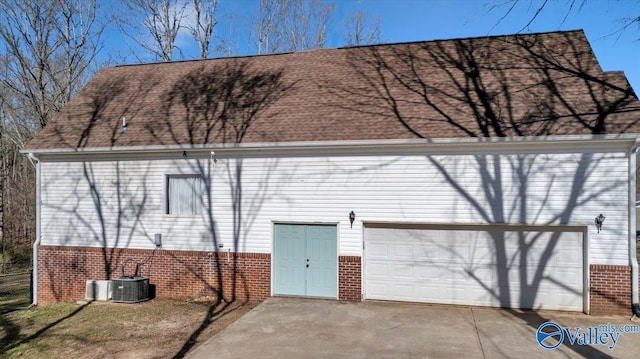 rear view of property with driveway, roof with shingles, central AC, and brick siding